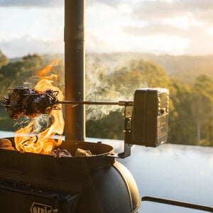 Outdoor Ozpig rotisserie grill with meat roasting at sunset.