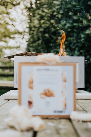 Blurry Kasai Wood Wool Firelighters box in foreground with fire burning in background, showcasing product in use.
