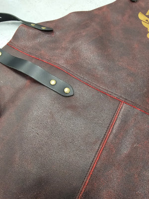 Close-up of Stalwart Crafts leather BBQ apron showing textured brown leather, red stitching, and black strap with brass rivets.