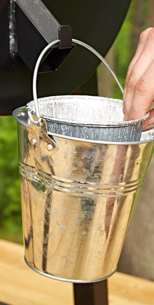A silver metal bucket with a wire handle and a foil pan inside, being held by a hand outdoors.