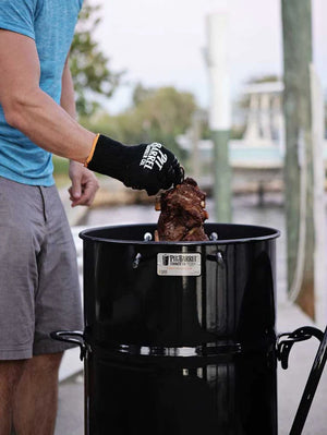 Griller wearing Pit Barrel Pit Gloves - FireFly Barbecue while handling meat in a Pit Barrel Cooker, perfect for safe and stylish BBQ handling.