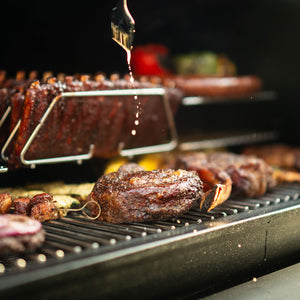 GMG Trek Cart with GMG Prime 2.0 RACKT Backboard in use, grilling steaks and ribs with stainless steel racks supporting even heat distribution.