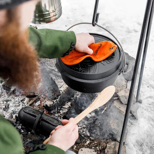 Person using Petromax 5.5L Cast Iron Dutch Oven over open fire with tripod setup, showcasing its outdoor cooking versatility and durability.