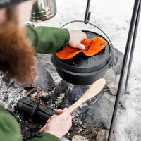 Person handling Petromax cast iron Dutch oven over campfire for outdoor cooking