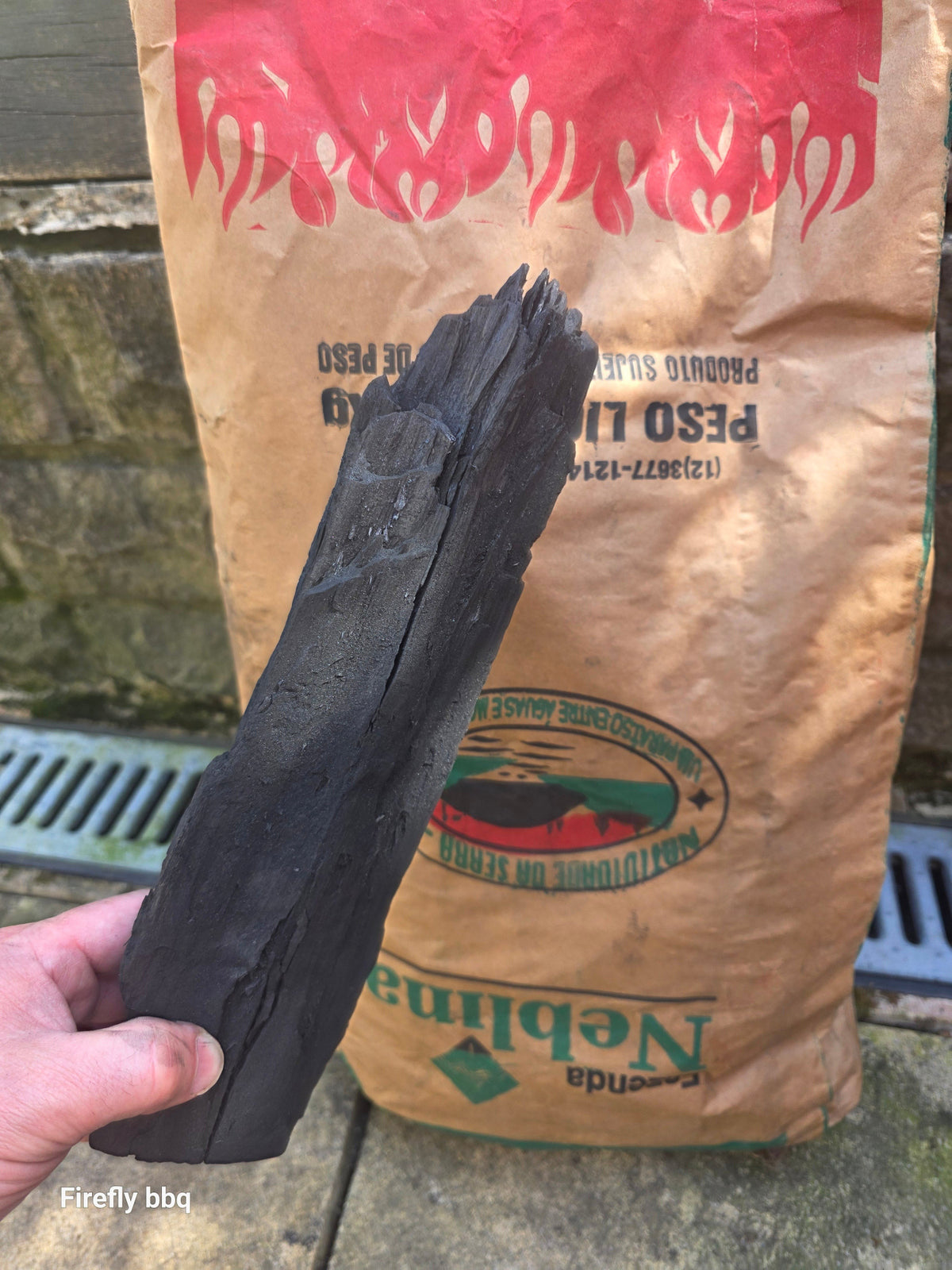A hand holding a dark charcoal briquette in front of a brown paper bag of Nebina brand barbecue coals, set against a stone wall background.