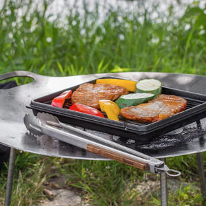 Petromax BBQ and Coal Tongs - Small resting beside grilled vegetables and meat on a fire bowl, ideal for outdoor kitchen and camping use.