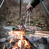 Person using BBQ tongs to handle food over campfire outdoors