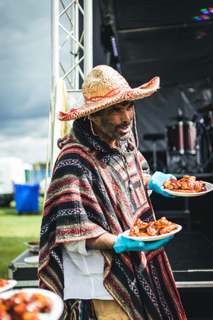 Man in poncho and sombrero serving spicy wings made with 7 Deadly Wings Hot Sauce at an outdoor food festival event.