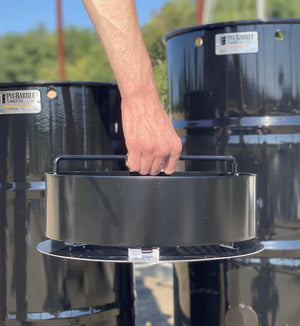 Hand lifting charcoal basket with FireFly Barbecue Pit Barrel Ash Catcher attached, demonstrating easy post-cook cleanup.