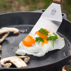 Petromax Flexible Spatula with Long Handle flipping sunny-side-up eggs in a cast iron Dutch oven, ideal for precise outdoor grilling tasks.