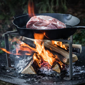 Petromax Cooking Stand supporting a cast iron Dutch oven with raw meat over a wood fire, ideal for outdoor grilling enthusiasts.