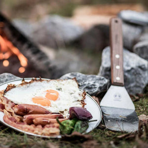 Petromax Flexible Spatula with Long Handle resting beside a plate of fried eggs and bacon, showcasing its use in outdoor Dutch oven cooking.