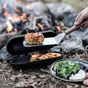 Petromax Flexible Spatula with Long Handle lifting grilled food from a Dutch oven over an open fire, perfect for outdoor cooking enthusiasts.