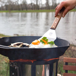 Petromax Flexible Spatula with Long Handle flipping eggs in a Dutch oven on a wood-fired grill, designed for outdoor kitchen and camping use.