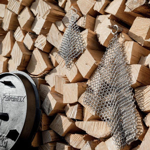 Two Petromax Chain Mail Cleaners hanging on stacked firewood, showcasing their rugged design for outdoor Dutch oven maintenance.