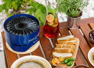 Cobalt blue Feuerhand Tamber Table Top Grill in use on a wooden table with food and drinks, perfect for outdoor dining and garden parties.
