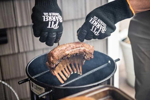 Man using Pit Barrel Pit Gloves - FireFly Barbecue to lift ribs from a smoker, showcasing heat protection and non-slip grip for outdoor cooking.