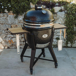 Monolith TWO 66 Kamado Grill in outdoor setting with closed lid, bamboo shelves, and grilling tools, designed for stylish and durable outdoor cooking.