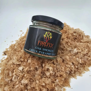 Firefly BBQ Oak Smoked Greek Pyramid Salt jar lying on a bed of golden smoked salt flakes, emphasizing artisanal quality.