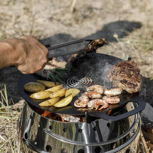 Petromax Atago grill plate cooking shrimp, steak, and potatoes over open flame, perfect for outdoor grilling enthusiasts.