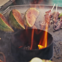 Outdoor grilling scene with sliced fruit, skewered meat, and flames on Kamado grill for outdoor cooking