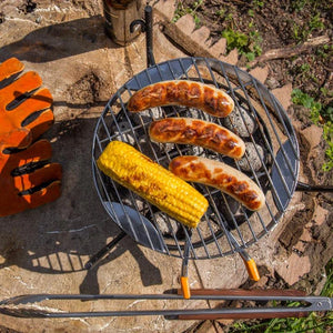Petromax Grilling Grate in use with sausages and corn on Atago grill, demonstrating outdoor cooking performance and heat distribution.