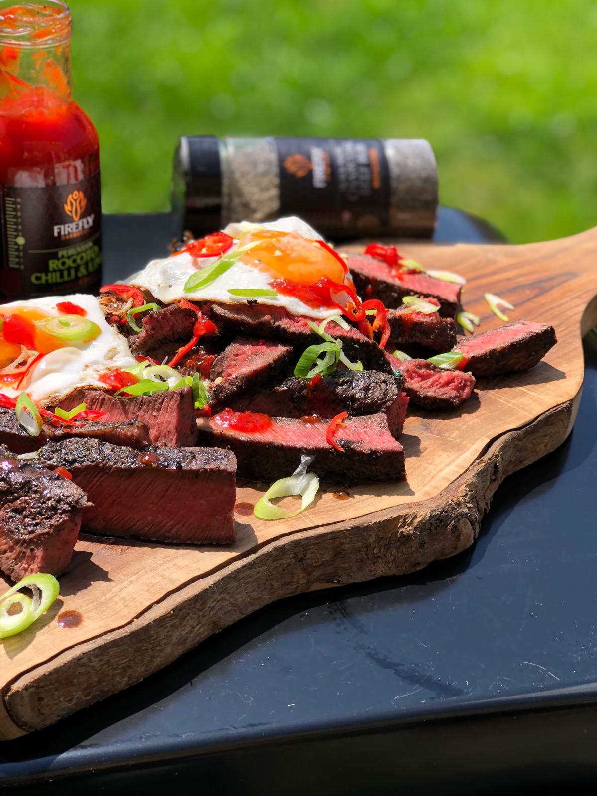 Grilled steak slices topped with Firefly BBQ Peruvian Rocoto Chilli & Lime Sauce, garnished with spring onions and fried eggs on wooden board.