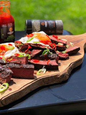 Grilled steak slices topped with Firefly BBQ Peruvian Rocoto Chilli & Lime Sauce, garnished with spring onions and fried eggs on wooden board.