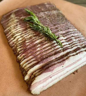 Sliced dry-cured bacon seasoned with rosemary and black pepper, showcasing the results of using the Sweet Rosemary & Thyme Bacon Cure for pork.