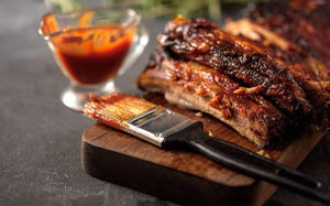 Barbecue ribs brushed with Kansas Brisket BBQ Sauce on a wooden board, served with sauce and brush for rich, smoky flavor presentation.