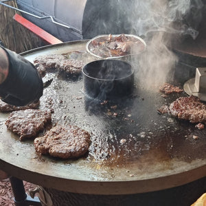 Monolith Fire Plate - LeCHEF in use with burgers and onions grilling on steel surface, ideal for kamado-style barbecue cooking.