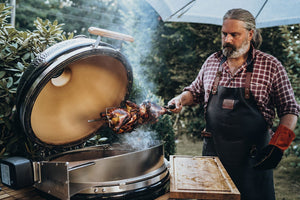 Man grilling rotisserie chicken on Monolith Le Chef Pro Series 2.0 Kamado Grill, showcasing its versatility for slow roasting outdoors.