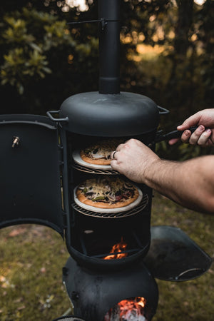 User placing pizzas inside OzPig Oven Smoker with visible flames below. Ideal for outdoor pizza making and multi-level cooking.
