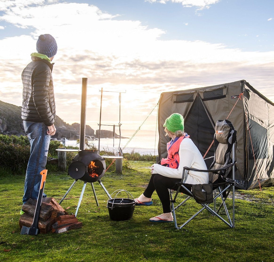 Couple using Ozpig Series 2 Portable Wood Fired BBQ Stove at a campsite near the ocean, highlighting its portability and outdoor cooking features.