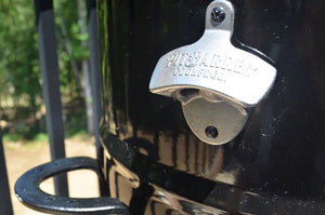 Close-up of Pit Barrel Bottle Opener - FireFly Barbecue mounted on a black outdoor grill, perfect for BBQ and beer lovers.