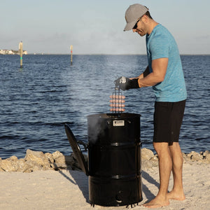Man grilling sausages on a beach using Pit Barrel Sausage, Hot Dog & Brat Hanger - FireFly Barbecue with a Pit Barrel Cooker.