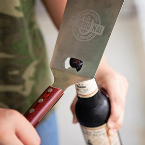 Pit Barrel Ultimate Spatula being used to open a beer bottle, highlighting its multifunctional design for outdoor cooking and entertaining.