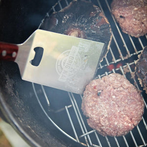 Pit Barrel Ultimate Spatula flipping burgers on a grill, designed for heavy-duty outdoor cooking with a built-in bottle opener and sturdy grip.