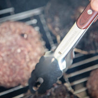 Close-up of Pit Barrel Ultimate Tongs flipping burgers on a grill, showing engraved branding and durable stainless steel construction.