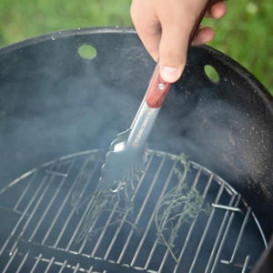 Using Pit Barrel Ultimate Tongs to place herbs on a grill, highlighting precision grip and heat-resistant wood handle for outdoor cooking.
